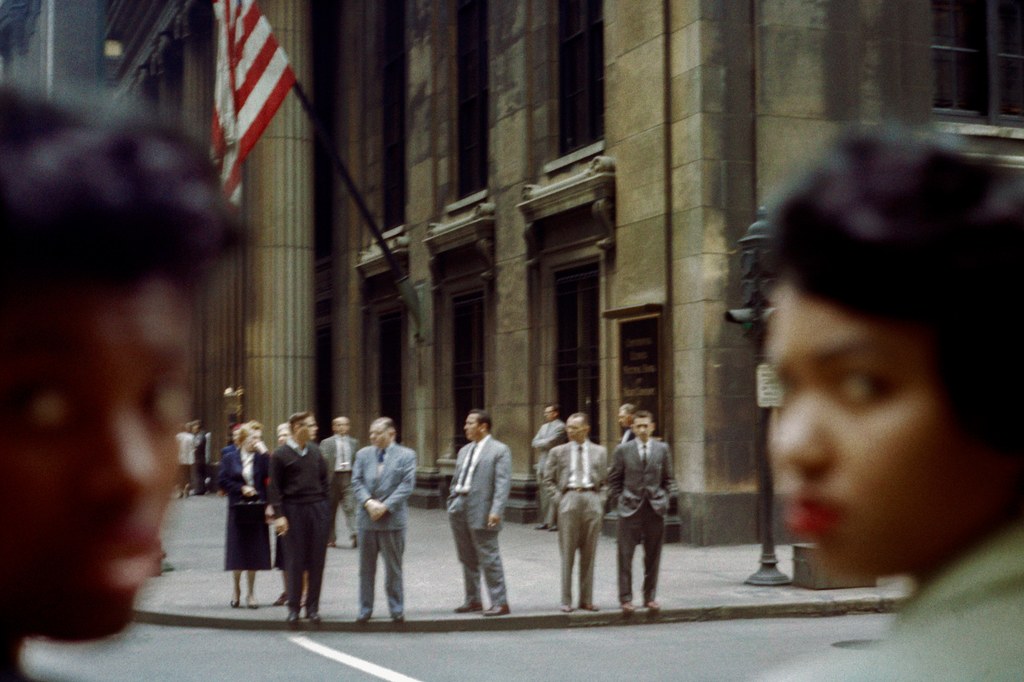 Photograph by Vivian Maier of two women on the sides of the frame are out of focus and looking at the camera. People stand in between their faces.
