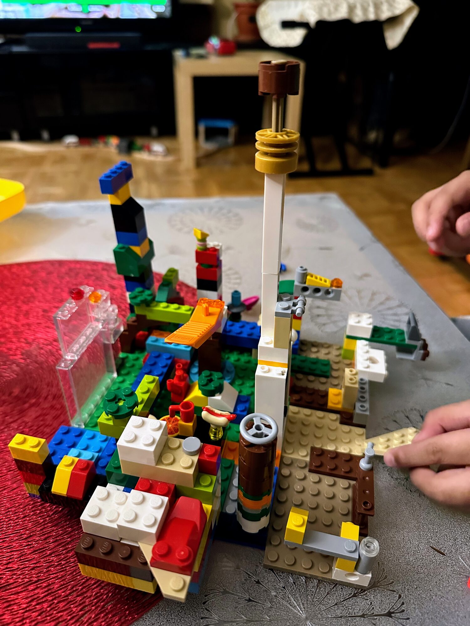 A child's hands adding a piece to a tall, sprawling Lego build of mismatched bricks, towers and platforms on a living-room table